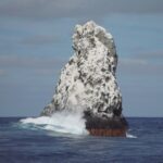 Il picco nord-ovest di Roca Partida, bianco di guano, visto dalla barca, con un'onda che si infrange alla base e uccelli in volo sopra