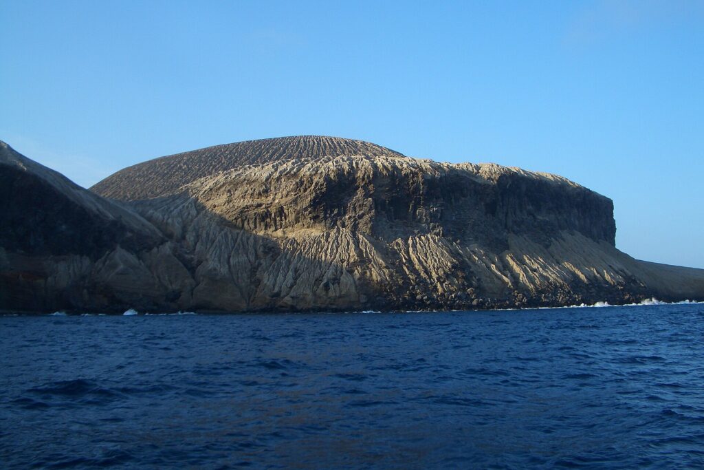 La costa di San Benedicto vista dal mare: il cono di cenere del vulcano Bárcena, solcato da canaloni, sopra scogliere scure e un mare blu scuro