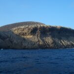 La costa di San Benedicto vista dal mare: il cono di cenere del vulcano Bárcena, solcato da canaloni, sopra scogliere scure e un mare blu scuro