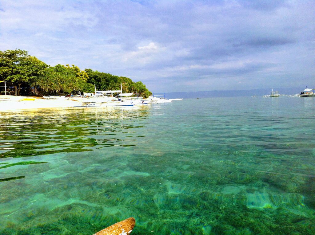 Acqua turchese trasparente sopra un fondale basso, davanti a una spiaggia di sabbia bianca orlata di alberi dove sono allineate bangka bianche, altre barche al largo sotto un cielo nuvoloso.