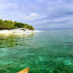 Acqua turchese trasparente sopra un fondale basso, davanti a una spiaggia di sabbia bianca orlata di alberi dove sono allineate bangka bianche, altre barche al largo sotto un cielo nuvoloso.