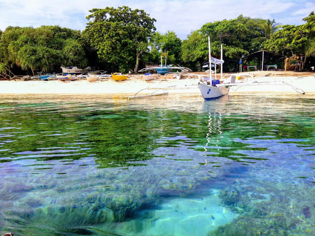 A filo d'acqua, blocchi di corallo sotto un'acqua chiara e turchese, davanti a una spiaggia di sabbia bianca dove è ancorata una bangka bianca, altre barche colorate tirate in secco sotto gli alberi.
