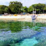 A filo d'acqua, blocchi di corallo sotto un'acqua chiara e turchese, davanti a una spiaggia di sabbia bianca dove è ancorata una bangka bianca, altre barche colorate tirate in secco sotto gli alberi.