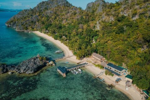 Vista aerea del Sangat Island Dive Resort, con la spiaggia, il pontile e le barche, ai piedi delle scogliere