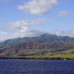 La costa dell'isola Socorro vista dal mare: falesie di lava nera, colline verdi e rossicce e la cima del vulcano tra le nuvole