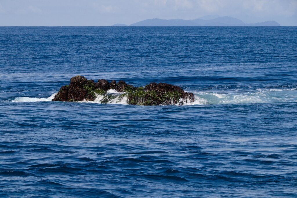 La cima di Richelieu Rock emerge con la bassa marea, una roccia scura coperta di alghe verdi battuta dalle onde, con le isole Surin all'orizzonte