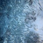 Un banco compatto di sardine argentate sfiora un fondale di sabbia chiarissima punteggiato di rocce scure, con la massa dei pesci più densa verso sinistra, nel blu.