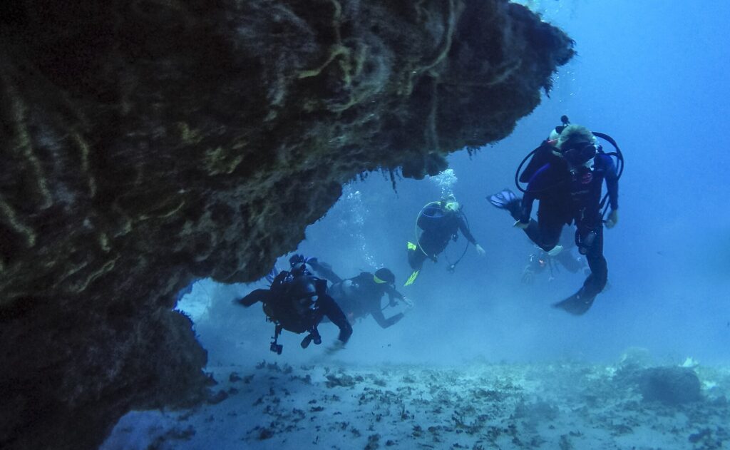 Quattro sub escono da un tunnel di corallo su un fondale di sabbia, a Cozumel, con la volta rocciosa sopra di loro