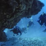 Quattro sub escono da un tunnel di corallo su un fondale di sabbia, a Cozumel, con la volta rocciosa sopra di loro