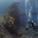 La prua sollevata del Giannis D, coperta di concrezioni, con tre sub ai piedi del relitto in un'acqua blu piena di bolle
