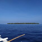 L'isola piatta e boscosa di Balicasag all'orizzonte, con il suo faro bianco e la frangia di sabbia chiara, vista da una bangka i cui bilancieri bianchi occupano il primo piano, sotto un cielo azzurro senza nuvole.