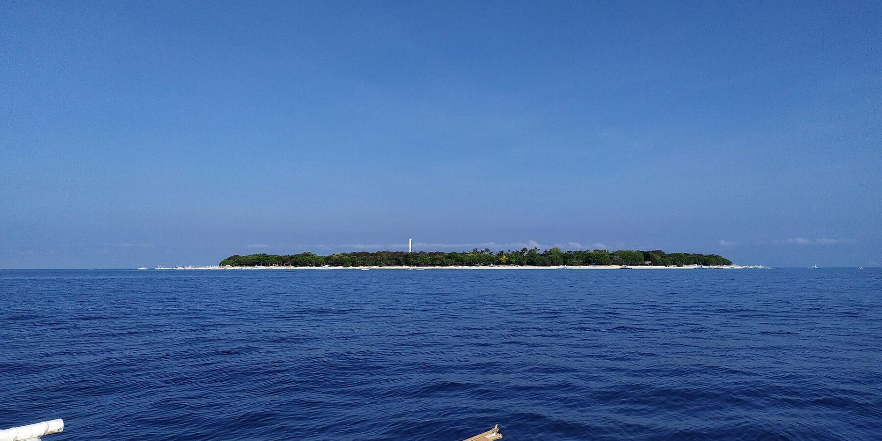 L'isola piatta e boscosa di Balicasag all'orizzonte, con il suo faro bianco e la frangia di sabbia chiara, vista da una bangka i cui bilancieri bianchi occupano il primo piano, sotto un cielo azzurro senza nuvole.