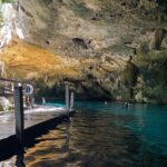 L'occhio est del cenote Dos Ojos: una volta di calcare irta di stalattiti sopra un'acqua turchese, un pontile di legno a sinistra e dei nuotatori sullo sfondo