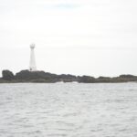 Un faro bianco slanciato con la porta rossa si erge sull'isolotto basso e roccioso di Formigão, rocce scure orlate di alghe brune, sopra un mare grigio e agitato sotto un cielo coperto