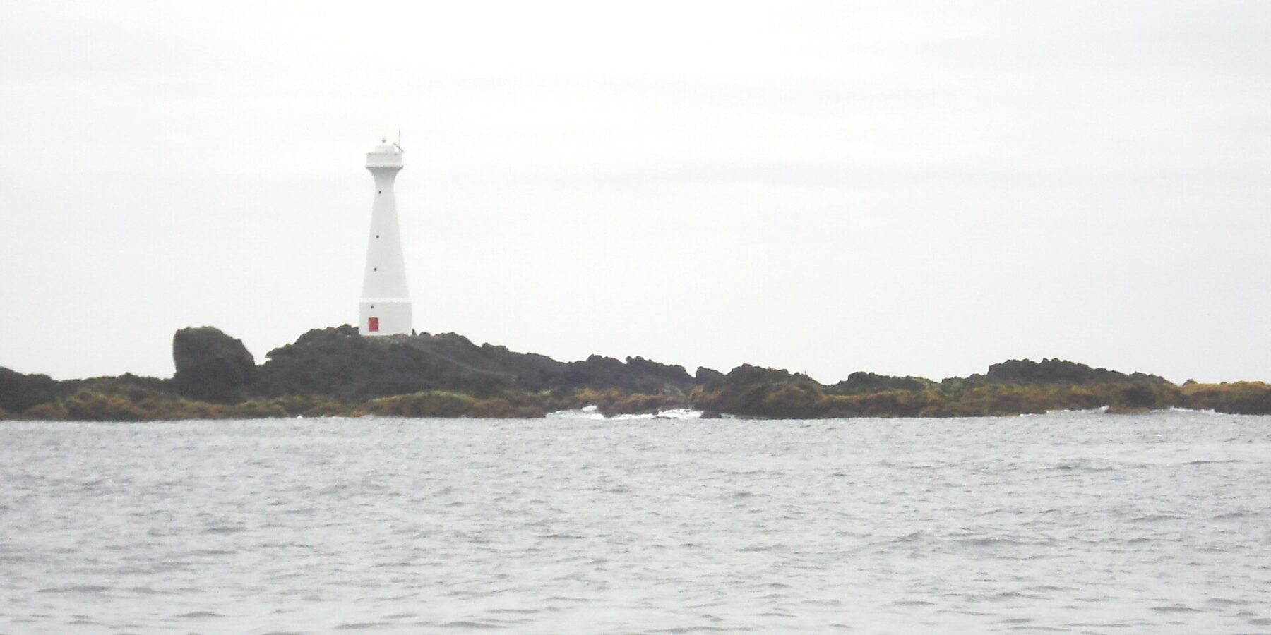 Un faro bianco slanciato con la porta rossa si erge sull'isolotto basso e roccioso di Formigão, rocce scure orlate di alghe brune, sopra un mare grigio e agitato sotto un cielo coperto