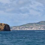 Panorama dei due isolotti di tufo ocra di Madalena su un mare blu scuro, l'Ilhéu Deitado lungo e piatto a sinistra e l'Ilhéu em Pé alto e dritto a destra, con la città bianca di Horta e le colline verdi di Faial sullo sfondo