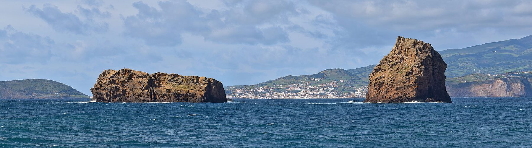 Panorama dei due isolotti di tufo ocra di Madalena su un mare blu scuro, l'Ilhéu Deitado lungo e piatto a sinistra e l'Ilhéu em Pé alto e dritto a destra, con la città bianca di Horta e le colline verdi di Faial sullo sfondo