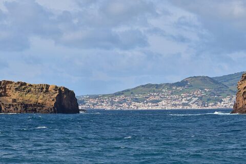 Panorama dei due isolotti di tufo ocra di Madalena su un mare blu scuro, l'Ilhéu Deitado lungo e piatto a sinistra e l'Ilhéu em Pé alto e dritto a destra, con la città bianca di Horta e le colline verdi di Faial sullo sfondo