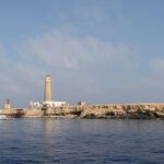Big Brother vista dal mare: un isolotto piatto e chiaro sormontato dal suo faro, con una barca da crociera bianca ormeggiata davanti