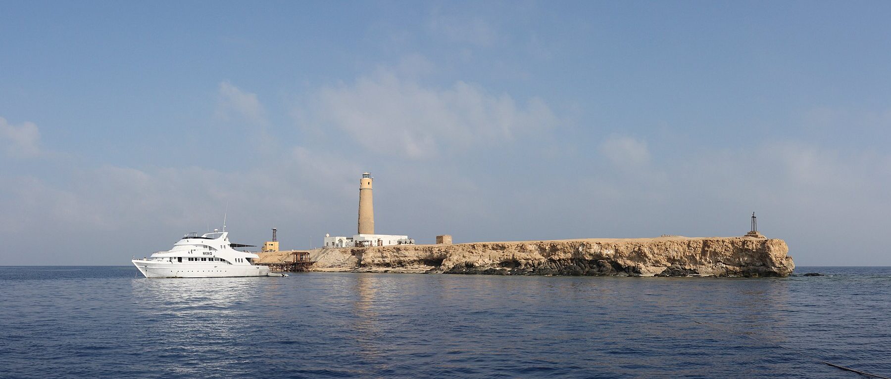 Big Brother vista dal mare: un isolotto piatto e chiaro sormontato dal suo faro, con una barca da crociera bianca ormeggiata davanti