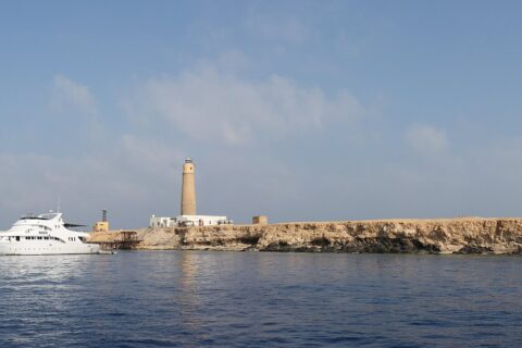 Big Brother vista dal mare: un isolotto piatto e chiaro sormontato dal suo faro, con una barca da crociera bianca ormeggiata davanti