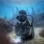 Un sub in muta nera, sdraiato su un fondale di sabbia tra due rocce coperte di alghe brune, scrive su una lavagnetta bianca, con le sagome delle laminarie che si stagliano nel blu dietro di lui.