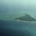 L'isola di Misali vista dall'aereo, un triangolo di foresta orlato da una spiaggia bianca, posato su un reef piatto turchese in mezzo a un mare blu scuro, nella foschia