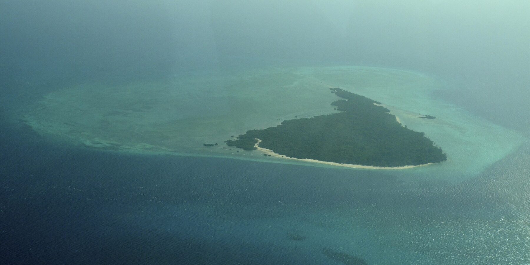 L'isola di Misali vista dall'aereo, un triangolo di foresta orlato da una spiaggia bianca, posato su un reef piatto turchese in mezzo a un mare blu scuro, nella foschia