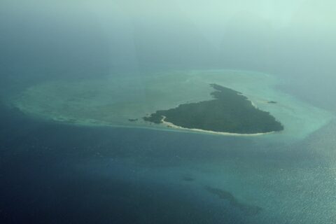 L'isola di Misali vista dall'aereo, un triangolo di foresta orlato da una spiaggia bianca, posato su un reef piatto turchese in mezzo a un mare blu scuro, nella foschia