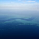 Vista dall'aereo di un canale blu scuro a forma di S che serpeggia tra le piattaforme turchesi della barriera corallina, con alcune piccole barche bianche nella passe