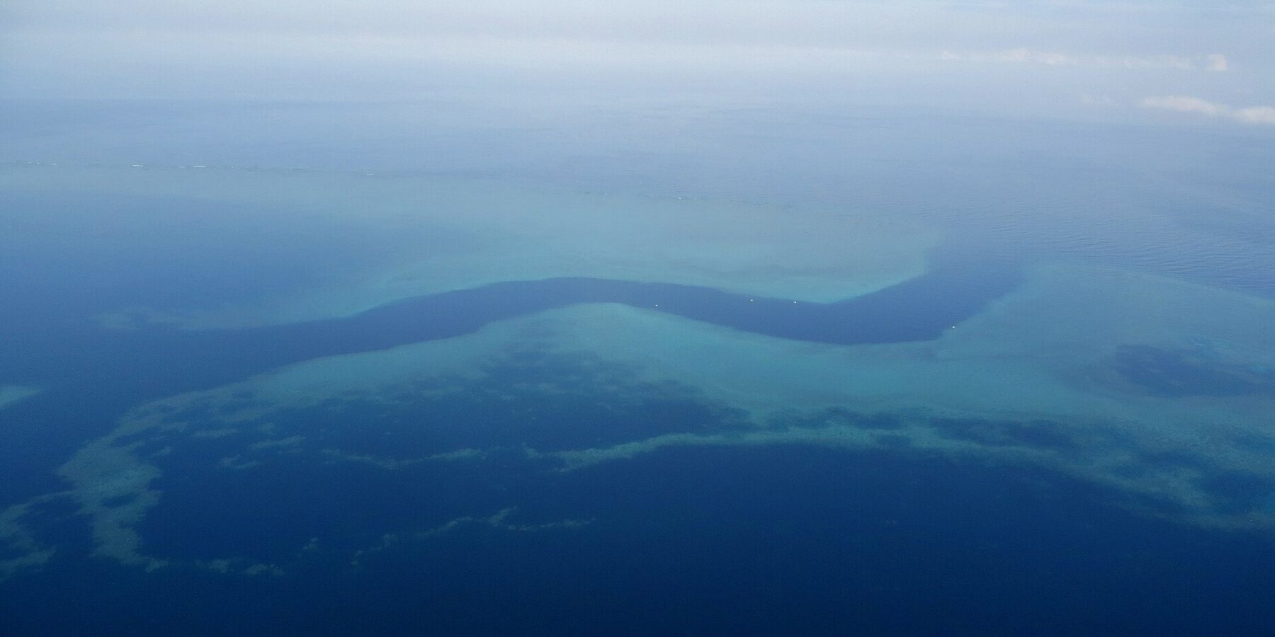 Vista dall'aereo di un canale blu scuro a forma di S che serpeggia tra le piattaforme turchesi della barriera corallina, con alcune piccole barche bianche nella passe