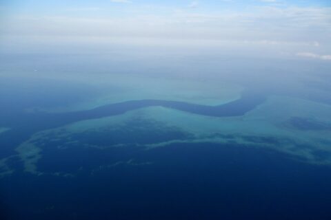 Vista dall'aereo di un canale blu scuro a forma di S che serpeggia tra le piattaforme turchesi della barriera corallina, con alcune piccole barche bianche nella passe
