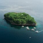 Vista aerea dell'isola di Pescador, uno scoglio coperto di vegetazione verde con il suo faro bianco, circondato da una decina di banca da immersione su un mare blu scuro