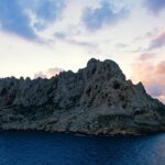 L'isola Maïre, massa di calcare chiaro dalle creste frastagliate, si staglia in controluce su un cielo al tramonto con nuvole rosa e malva, sopra un mare blu scuro.