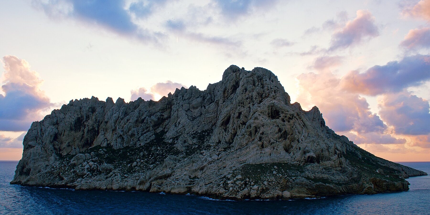 L'isola Maïre, massa di calcare chiaro dalle creste frastagliate, si staglia in controluce su un cielo al tramonto con nuvole rosa e malva, sopra un mare blu scuro.