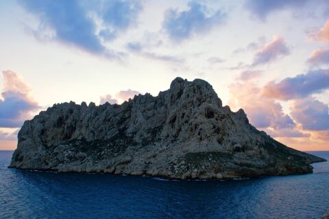 L'isola Maïre, massa di calcare chiaro dalle creste frastagliate, si staglia in controluce su un cielo al tramonto con nuvole rosa e malva, sopra un mare blu scuro.