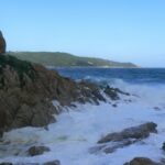 Onde che si infrangono in schiuma bianca sugli scogli di granito dell'Escalet, in primo piano, davanti a un mare blu scuro agitato, con in lontananza la costa boscosa di capo Camarat e il suo faro bianco.
