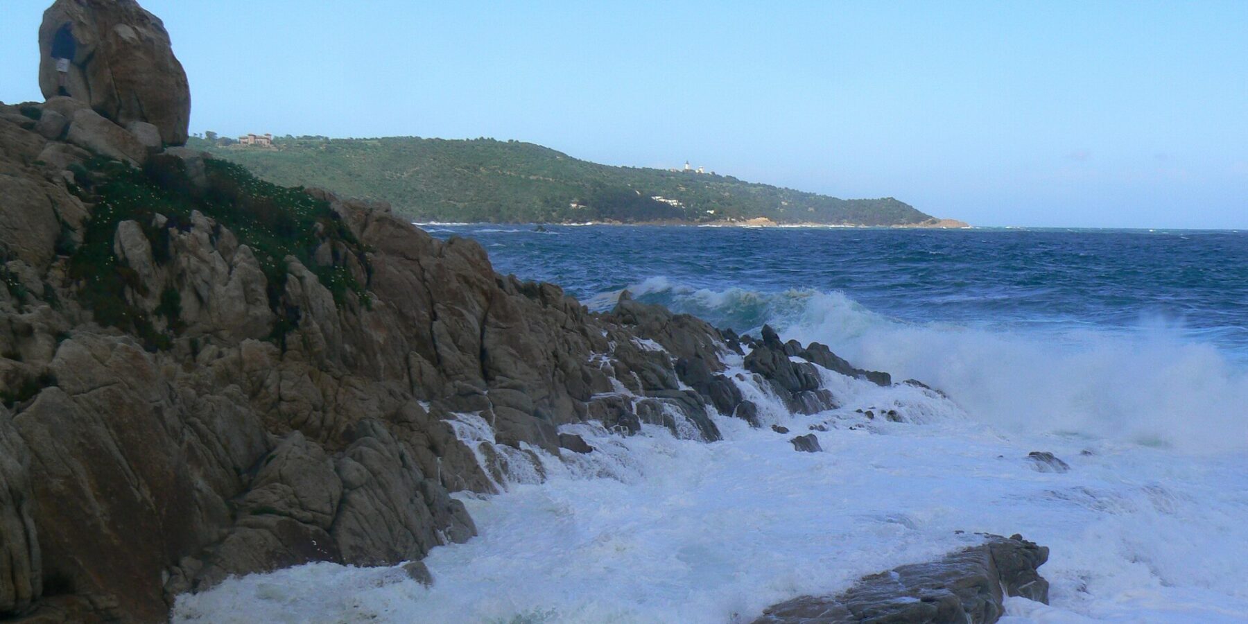 Onde che si infrangono in schiuma bianca sugli scogli di granito dell'Escalet, in primo piano, davanti a un mare blu scuro agitato, con in lontananza la costa boscosa di capo Camarat e il suo faro bianco.