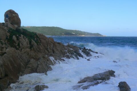 Onde che si infrangono in schiuma bianca sugli scogli di granito dell'Escalet, in primo piano, davanti a un mare blu scuro agitato, con in lontananza la costa boscosa di capo Camarat e il suo faro bianco.