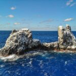 Roca Partida vista dal drone: uno scoglio imbiancato dagli uccelli, spaccato in due picchi, battuto da un mare blu scuro sotto un cielo azzurro con qualche nuvola