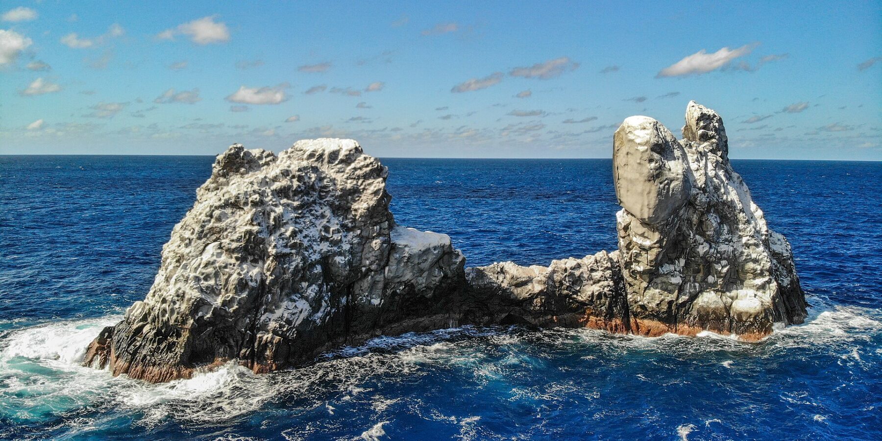 Roca Partida vista dal drone: uno scoglio imbiancato dagli uccelli, spaccato in due picchi, battuto da un mare blu scuro sotto un cielo azzurro con qualche nuvola