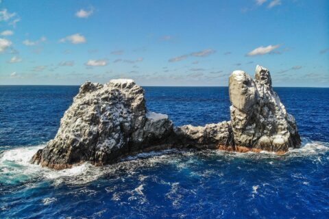 Roca Partida vista dal drone: uno scoglio imbiancato dagli uccelli, spaccato in due picchi, battuto da un mare blu scuro sotto un cielo azzurro con qualche nuvola