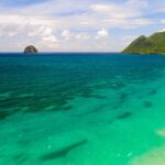 Vista aerea della lunga spiaggia di Le Diamant bordata di acqua turchese, con il Rocher du Diamant al largo e il morne Larcher, una collina verde, a destra