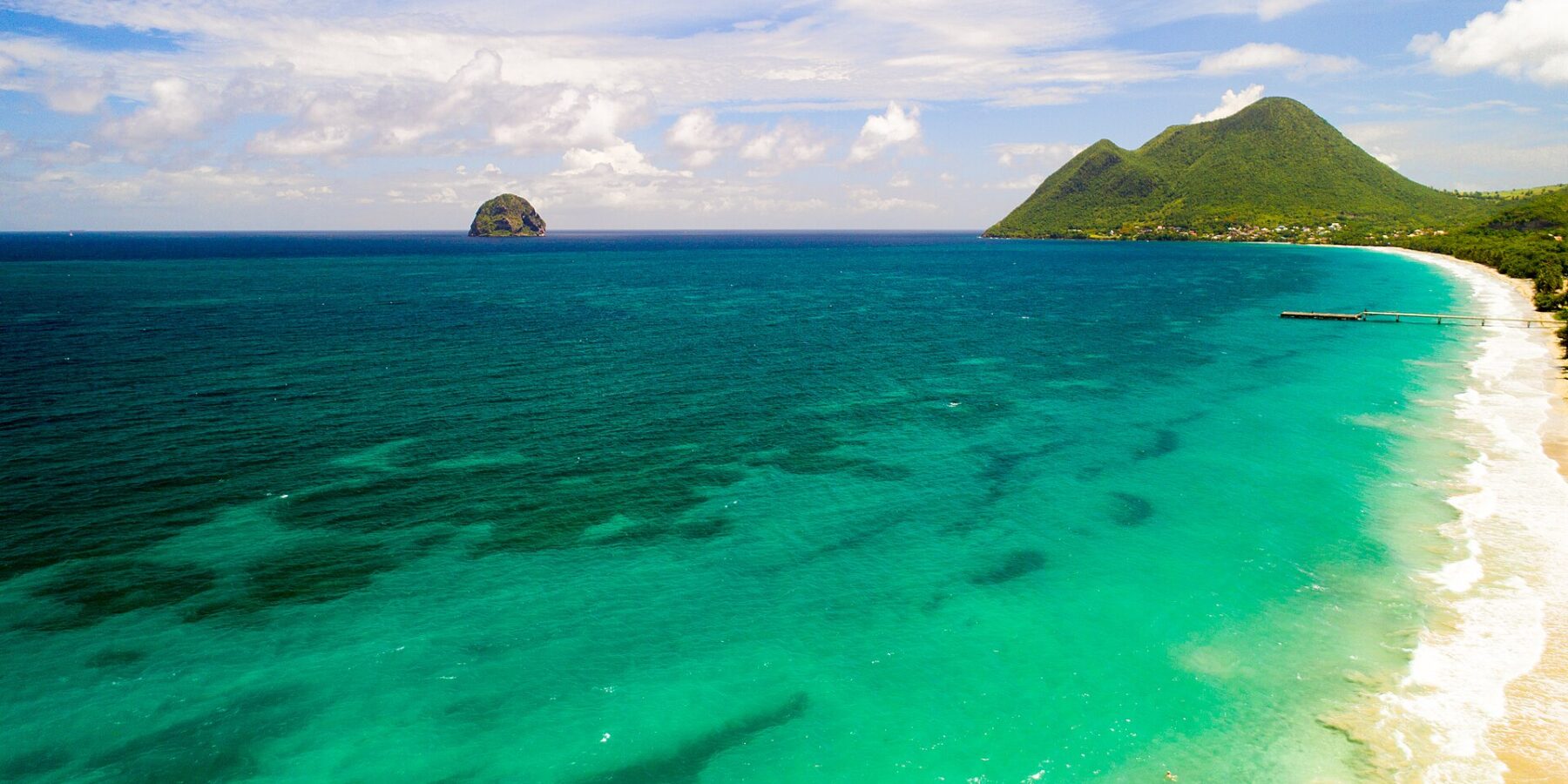 Vista aerea della lunga spiaggia di Le Diamant bordata di acqua turchese, con il Rocher du Diamant al largo e il morne Larcher, una collina verde, a destra