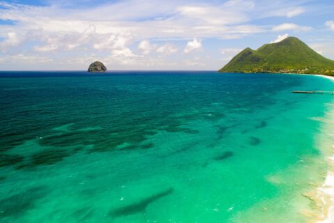 Vista aerea della lunga spiaggia di Le Diamant bordata di acqua turchese, con il Rocher du Diamant al largo e il morne Larcher, una collina verde, a destra