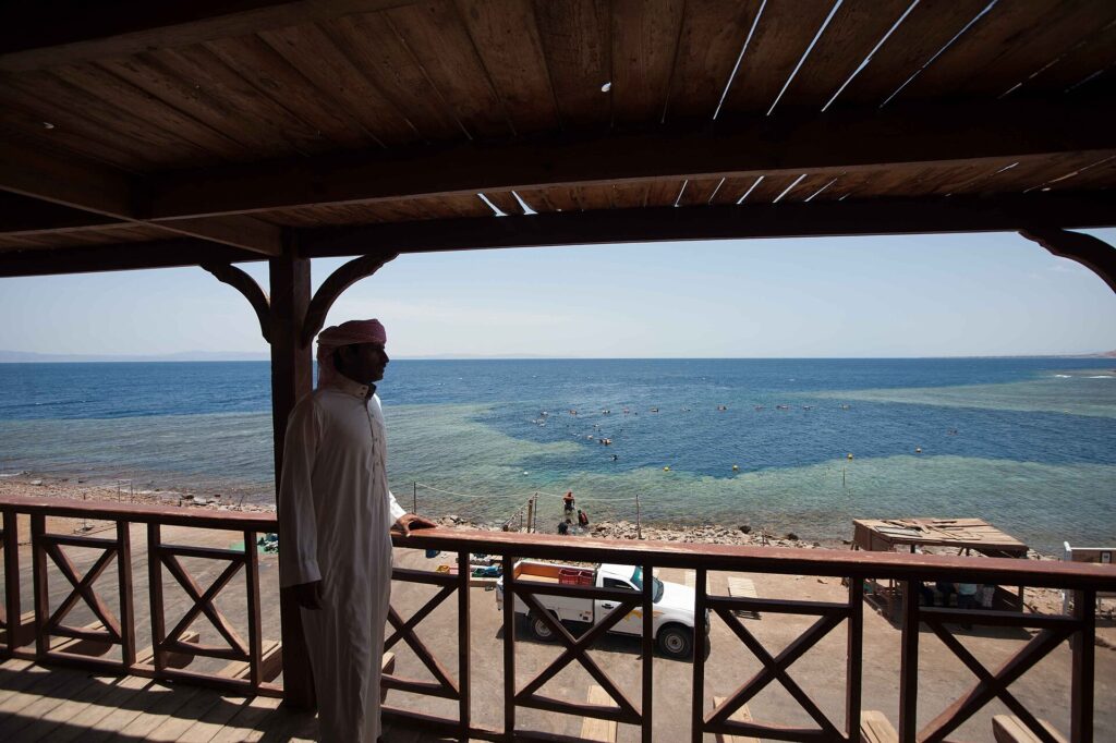 Da una terrazza di legno, un uomo in galabeya guarda la vasca del Blue Hole, orlata di reef chiaro, dove nuotano dei bagnanti