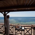 Da una terrazza di legno, un uomo in galabeya guarda la vasca del Blue Hole, orlata di reef chiaro, dove nuotano dei bagnanti