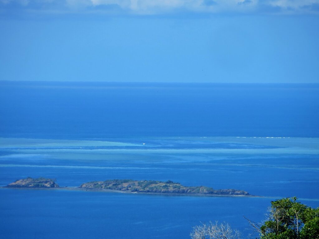 Vista dall'alto sulla laguna blu, due isolotti rocciosi in primo piano e, in lontananza, le piattaforme chiare della barriera corallina dove una barca bianca sosta nella passe