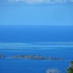 Vista dall'alto sulla laguna blu, due isolotti rocciosi in primo piano e, in lontananza, le piattaforme chiare della barriera corallina dove una barca bianca sosta nella passe
